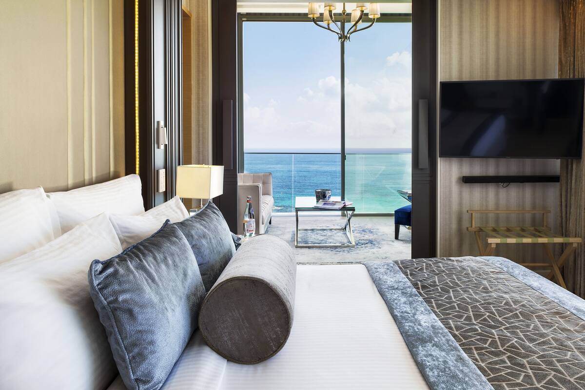 A hotel room with a bed, a TV, a table with a lamp, and a view of the ocean.