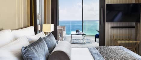 A hotel room with a bed, a TV, a table with a lamp, and a view of the ocean.