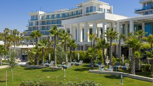 A modern hotel with multiple floors, large windows, and a well-maintained outdoor lounge area with palm trees and sun loungers. - Amavi, MadeForTwo Hotels - Paphos (Paphos)
