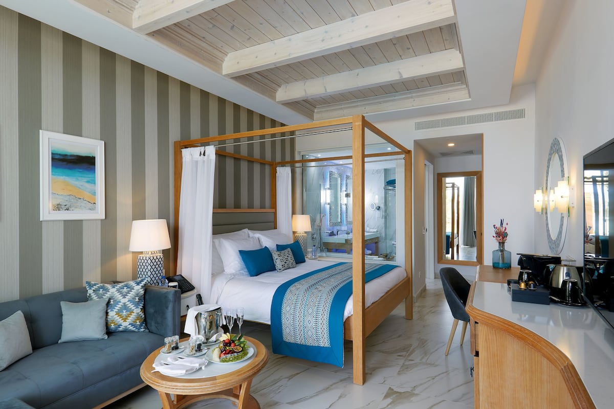A modern hotel room with a four-poster bed, a sitting area with a sofa and a small round table, a desk with a chair, and a large mirror.