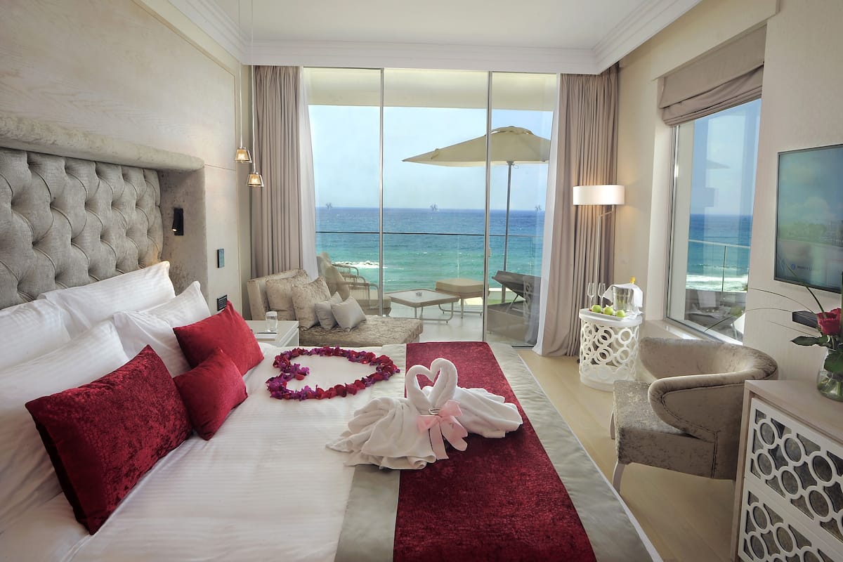 A hotel room with a large bed, a balcony with a view of the ocean, a TV, and a seating area with a table and chairs.