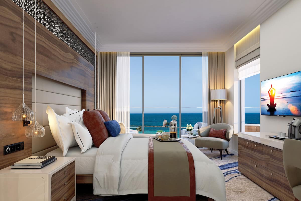 A modern hotel room with a large bed, a TV, a desk, and a view of the ocean.