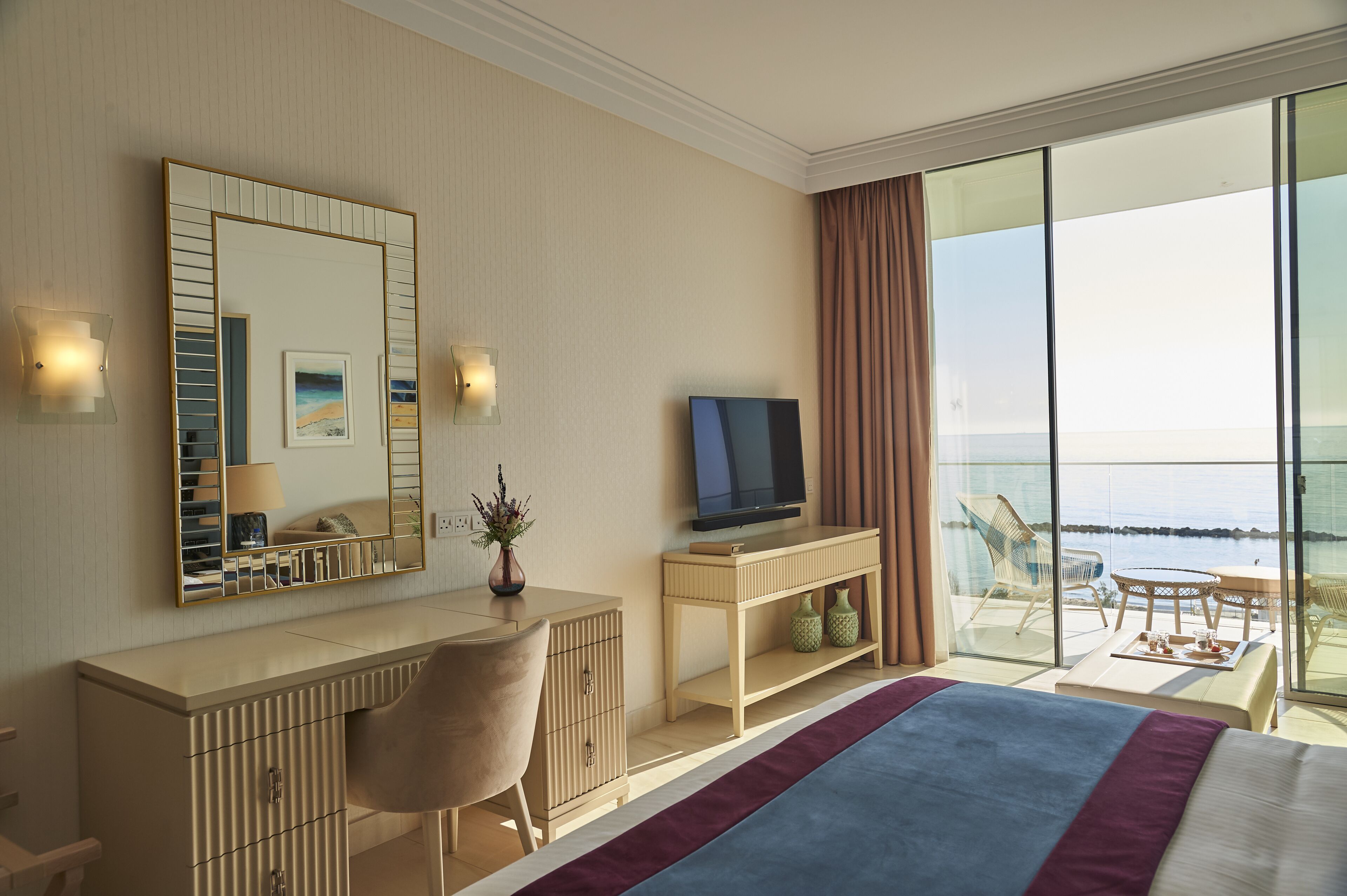 A hotel room with a bed, a desk, a television, a large mirror, and a balcony with a view of the sea.