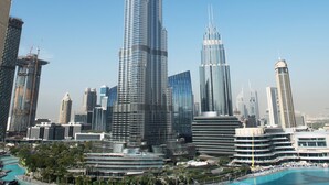 Un gratte-ciel moderne à la façade élégante et réfléchissante, entouré d’autres immeubles de grande hauteur et agrémenté, au premier plan, d’une grande fontaine circulaire.