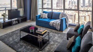 Un salon moderne avec un canapé bleu, des fauteuils gris et une table basse noire. Un téléviseur à écran plat est posé sur un meuble noir, et une grande fenêtre offre une vue sur la ville.