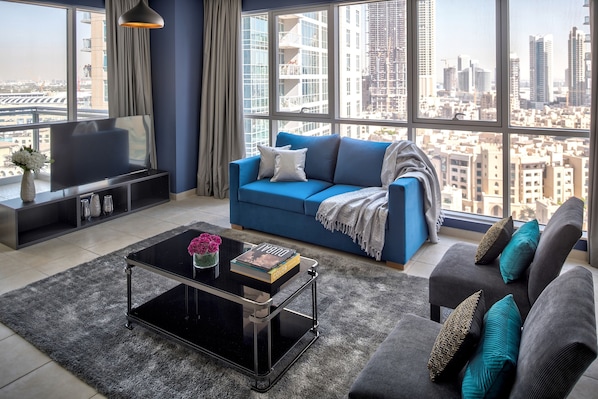 A modern living room with a blue sofa, grey armchairs, and a black coffee table. A flat-screen TV is on a black stand, and a large window offers a city view.