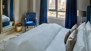 Une chambre d’hôtel avec un grand lit, un fauteuil bleu, une vue sur la ville par la fenêtre et un grand miroir.