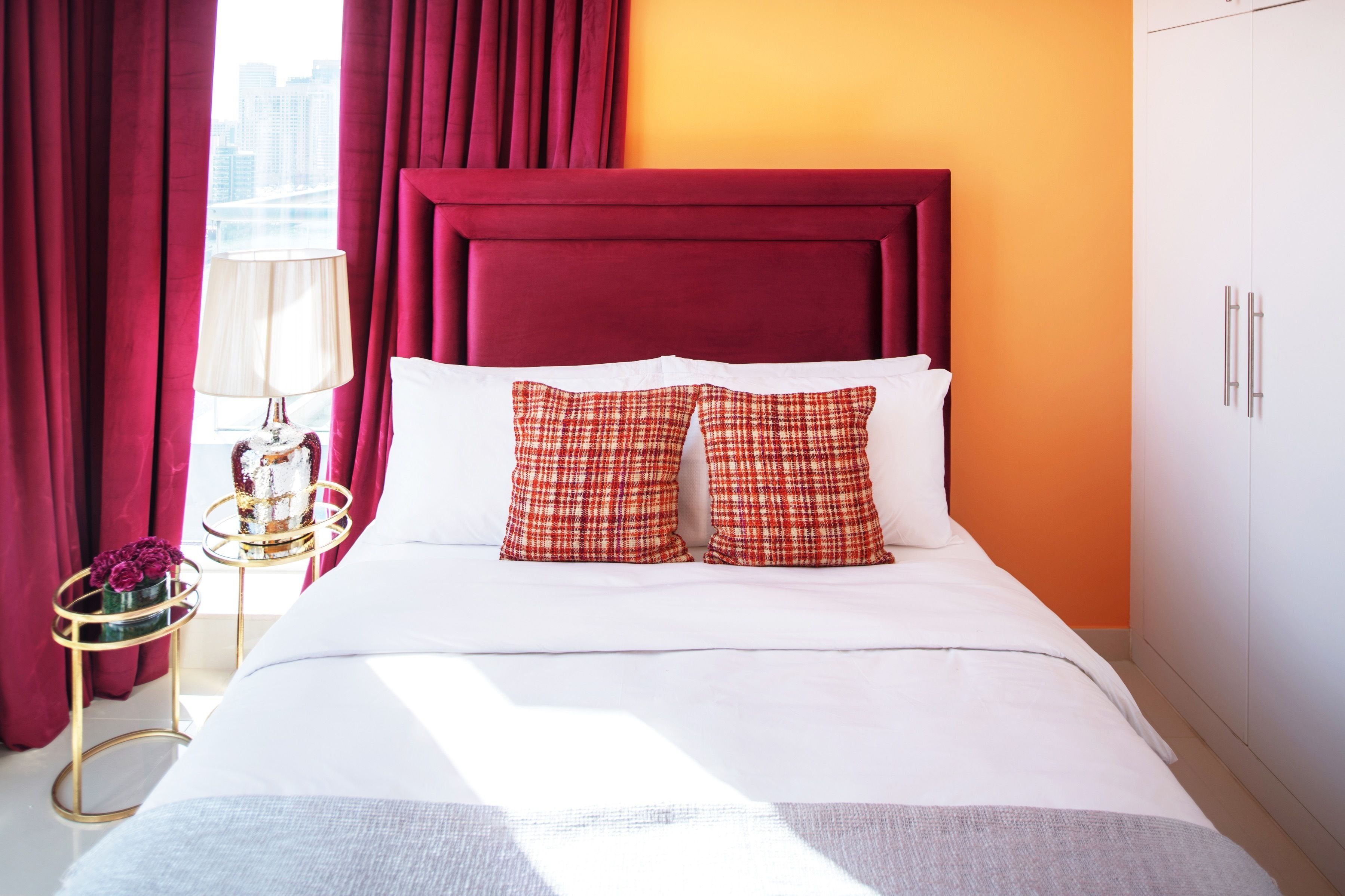 Quarto de hotel com uma cama grande, cortinas vermelhas, uma luminária e vista para a cidade.