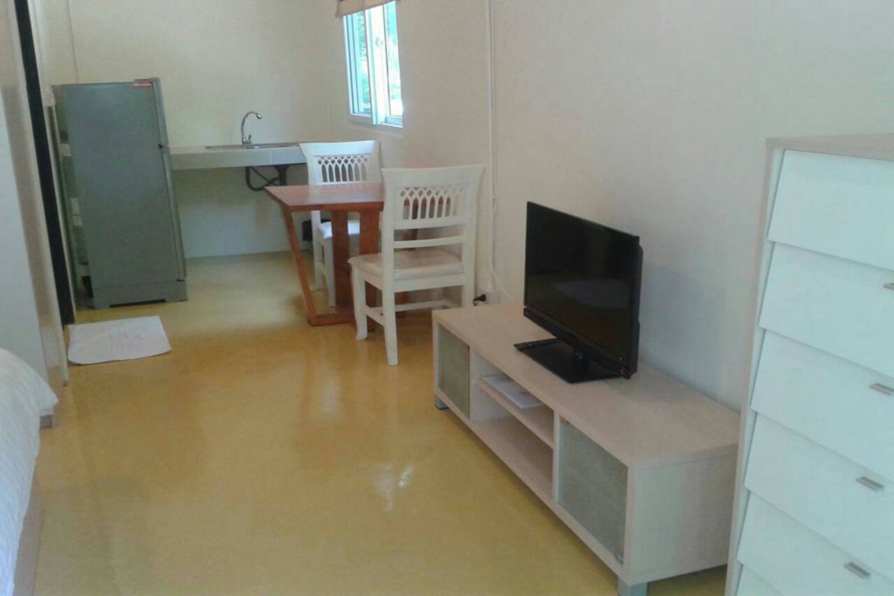 A compact room with a television on a stand, a small dining table with chairs, a refrigerator, and a window providing natural light.