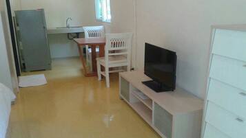 A compact room with a television on a stand, a small dining table with chairs, a refrigerator, and a window providing natural light.