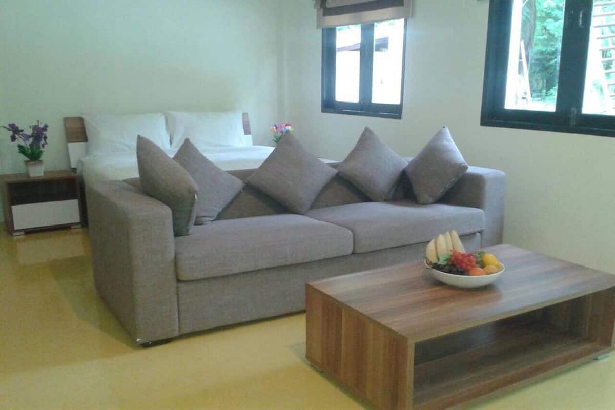 A modern living room with a grey sofa, a wooden coffee table with fruit, a bedside table with a lamp, and a window with a view of greenery.