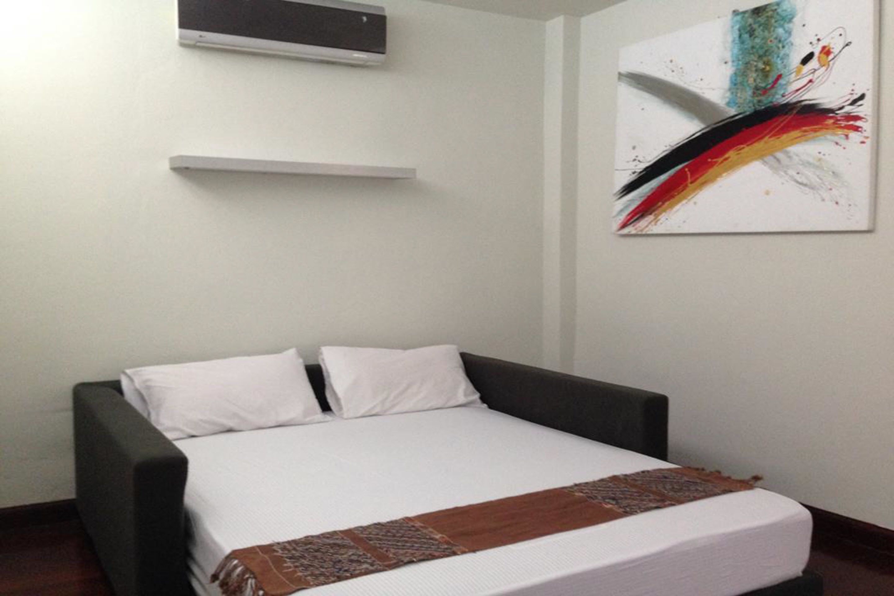 A bedroom with a bed, white pillows, a brown blanket, a white wall, and a colorful abstract painting.