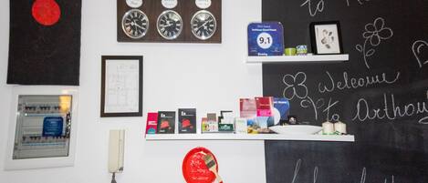Zona de recepción con una pizarra que muestra 'Welcome to Room 9' y 'Life is better'. Hay relojes, un teléfono y un certificado enmarcado en la pared.