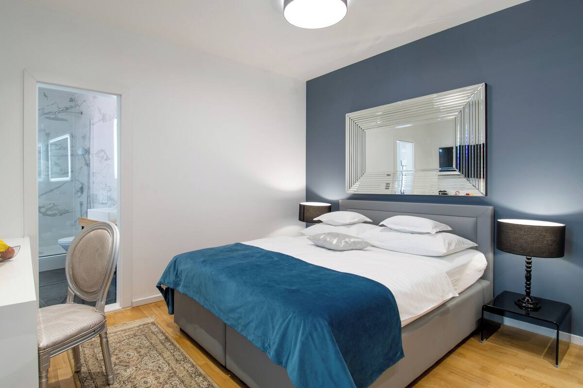A modern bedroom with a large bed, bedside lamps, a mirror, and a chair.