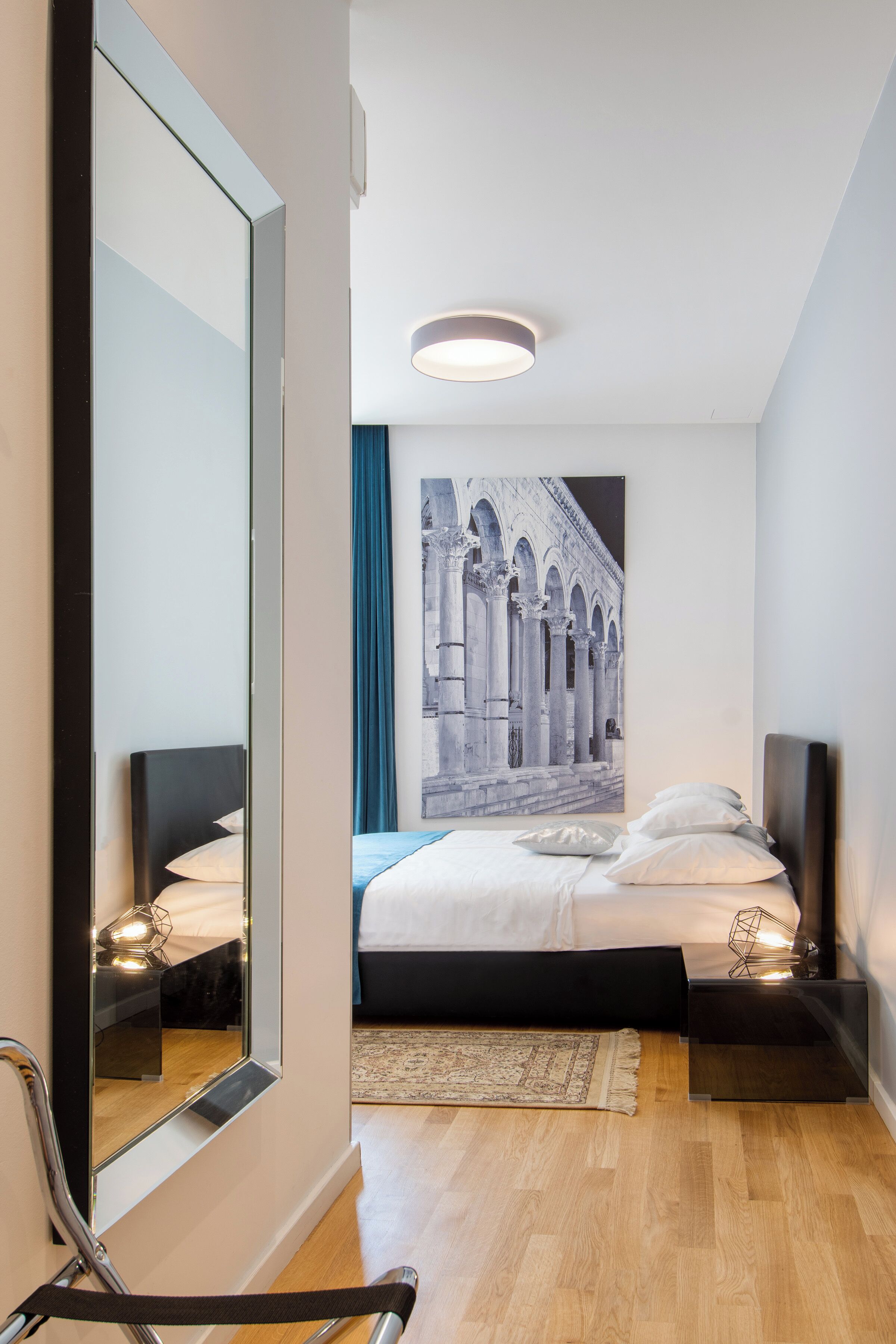 A modern hotel room with a large bed, a mirror, a nightstand, and a wall-mounted light fixture.