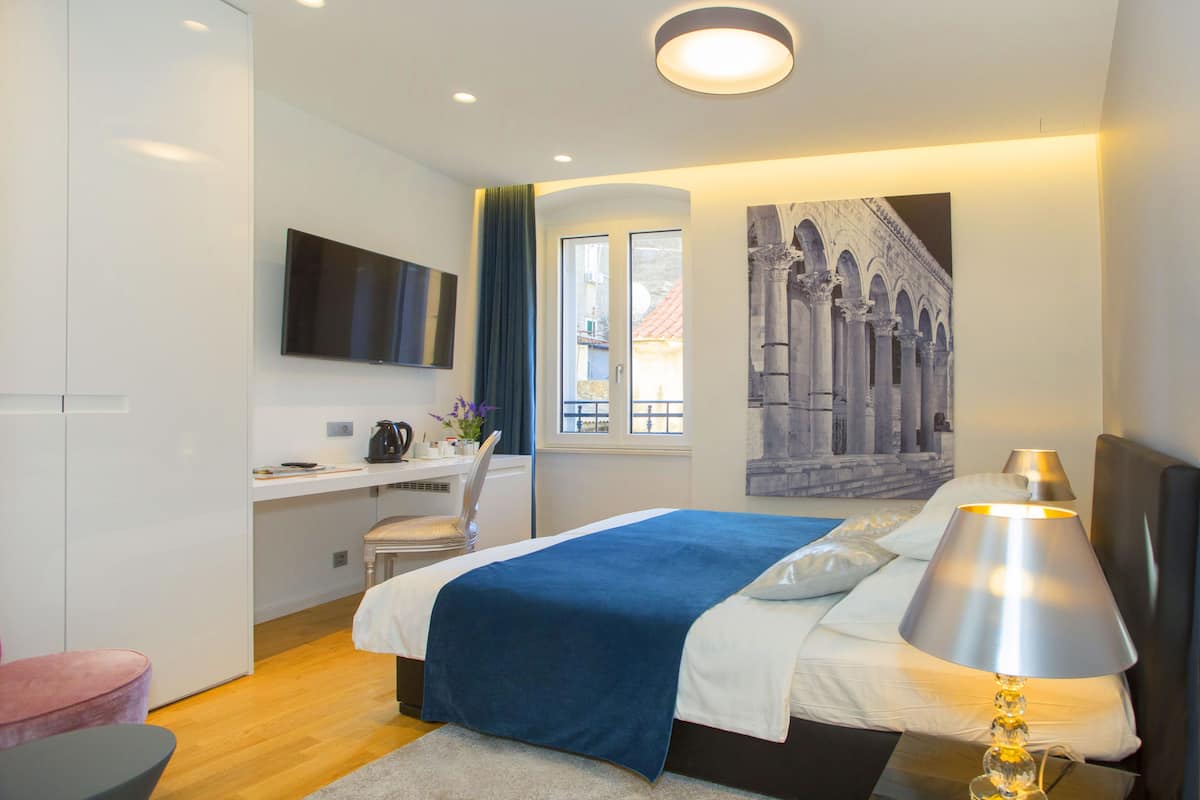 A modern bedroom with a bed, bedside lamp, desk, chair, and a large window.