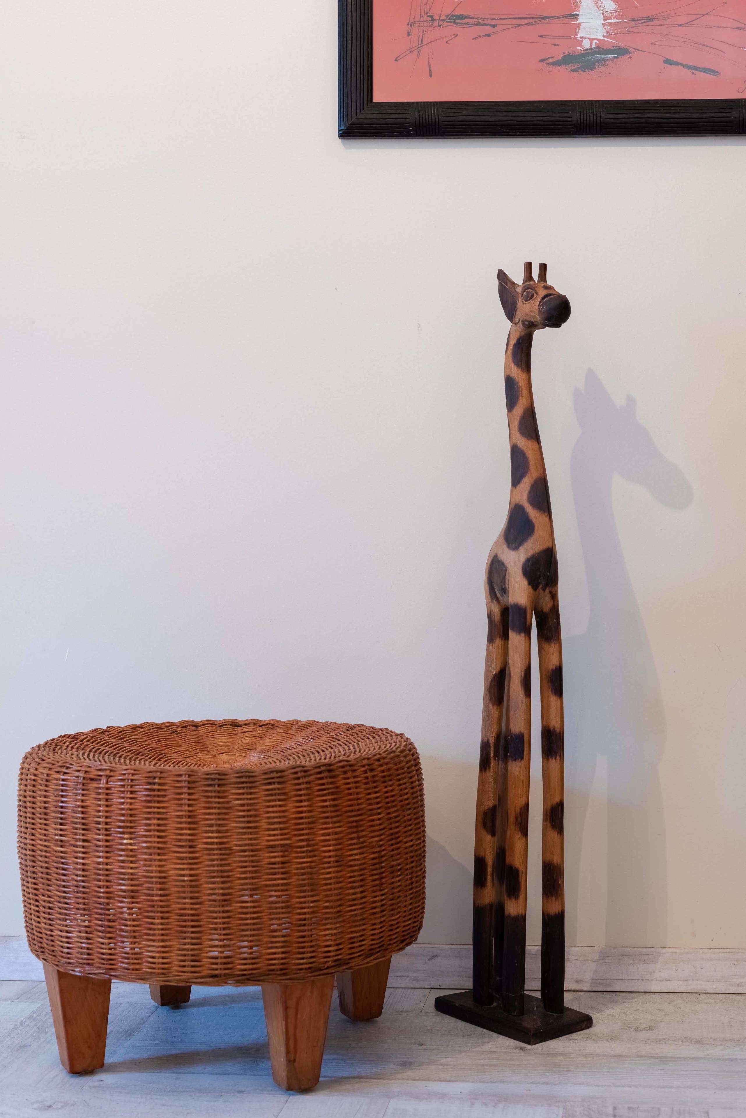 A wicker ottoman and a giraffe sculpture against a white wall with a framed picture above.