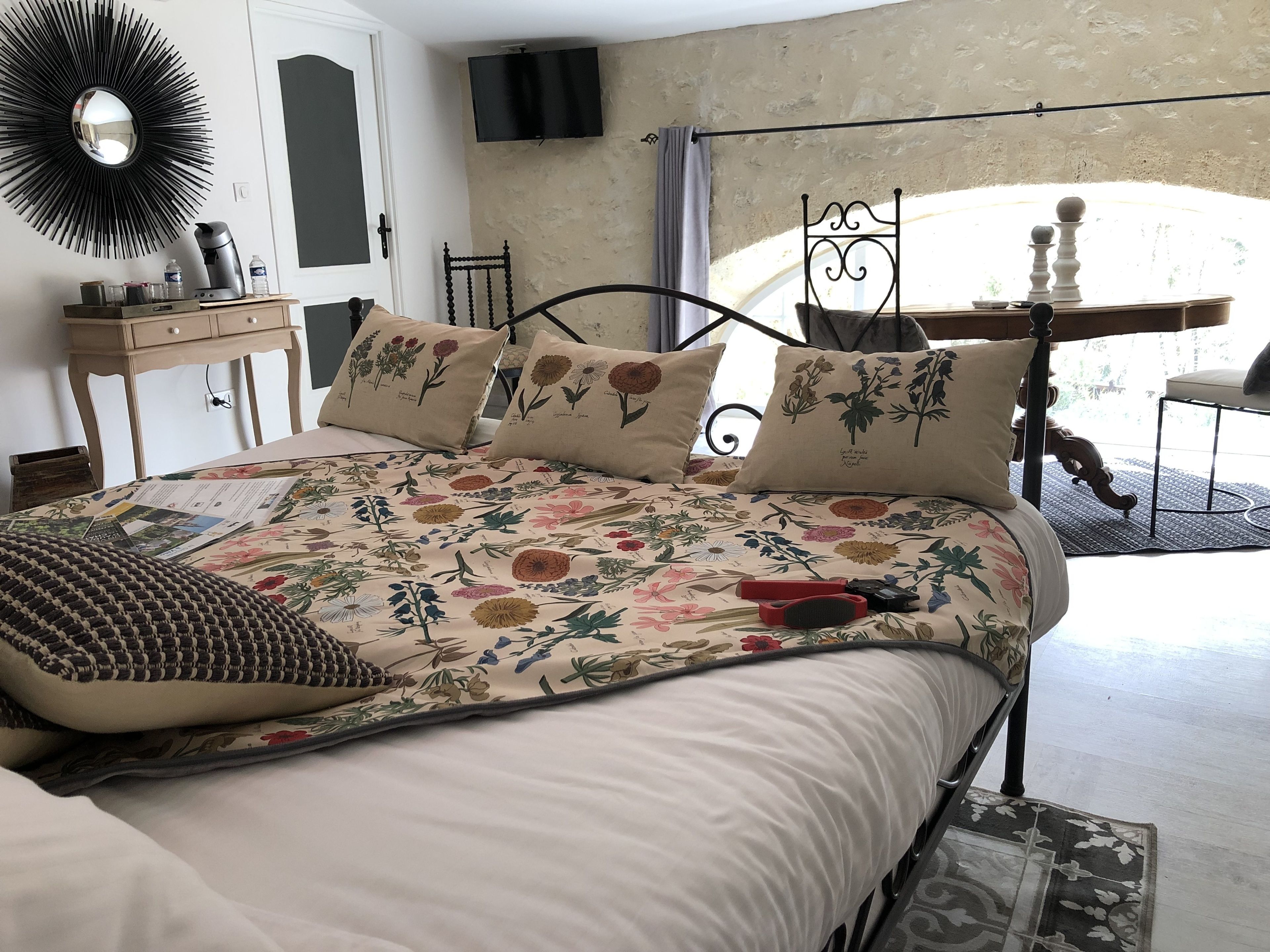 A bedroom with a floral-patterned bedspread, a round mirror, a television on the wall, and a view of the outdoors through a window.