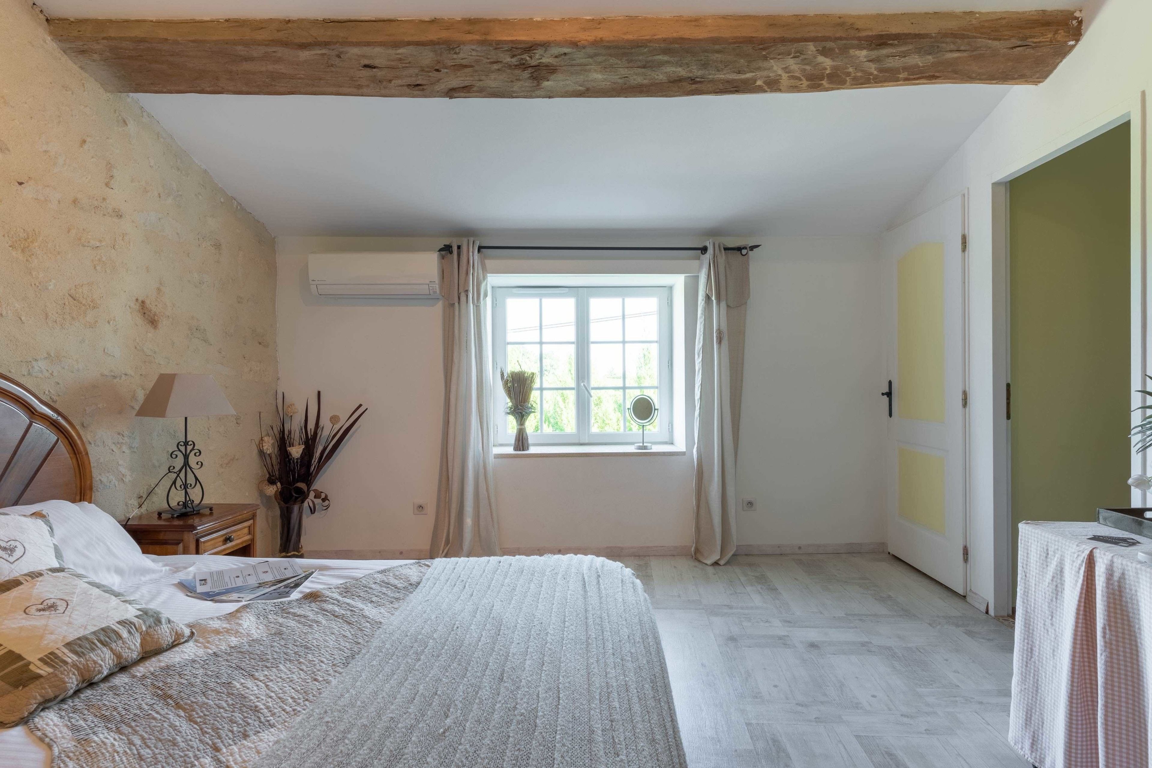 A bedroom with a bed, a window with curtains, a bedside table with a lamp, and a wall-mounted air conditioner.