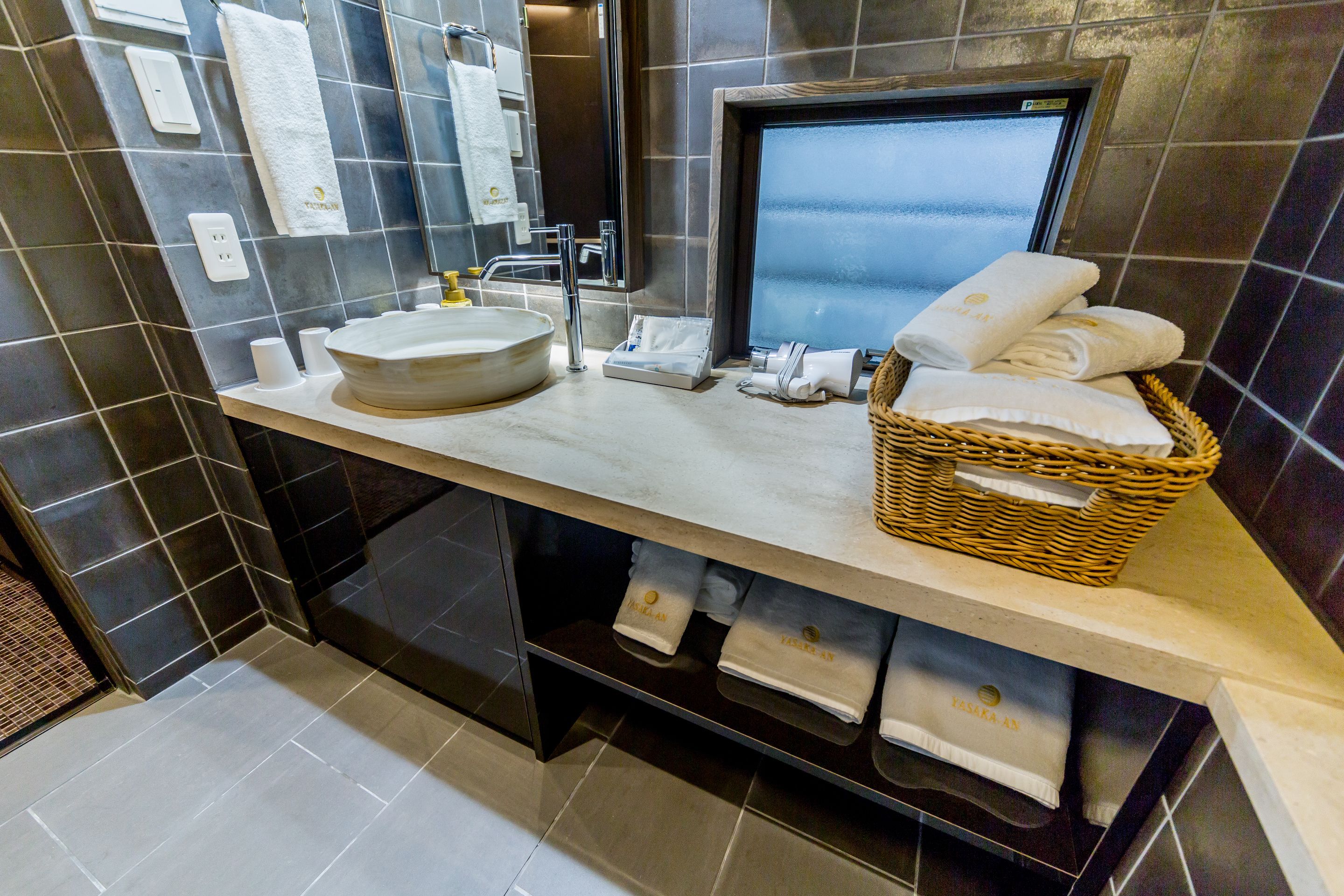 A modern bathroom with a large sink, mirror, and a digital screen. There are towels and a basket on the counter.