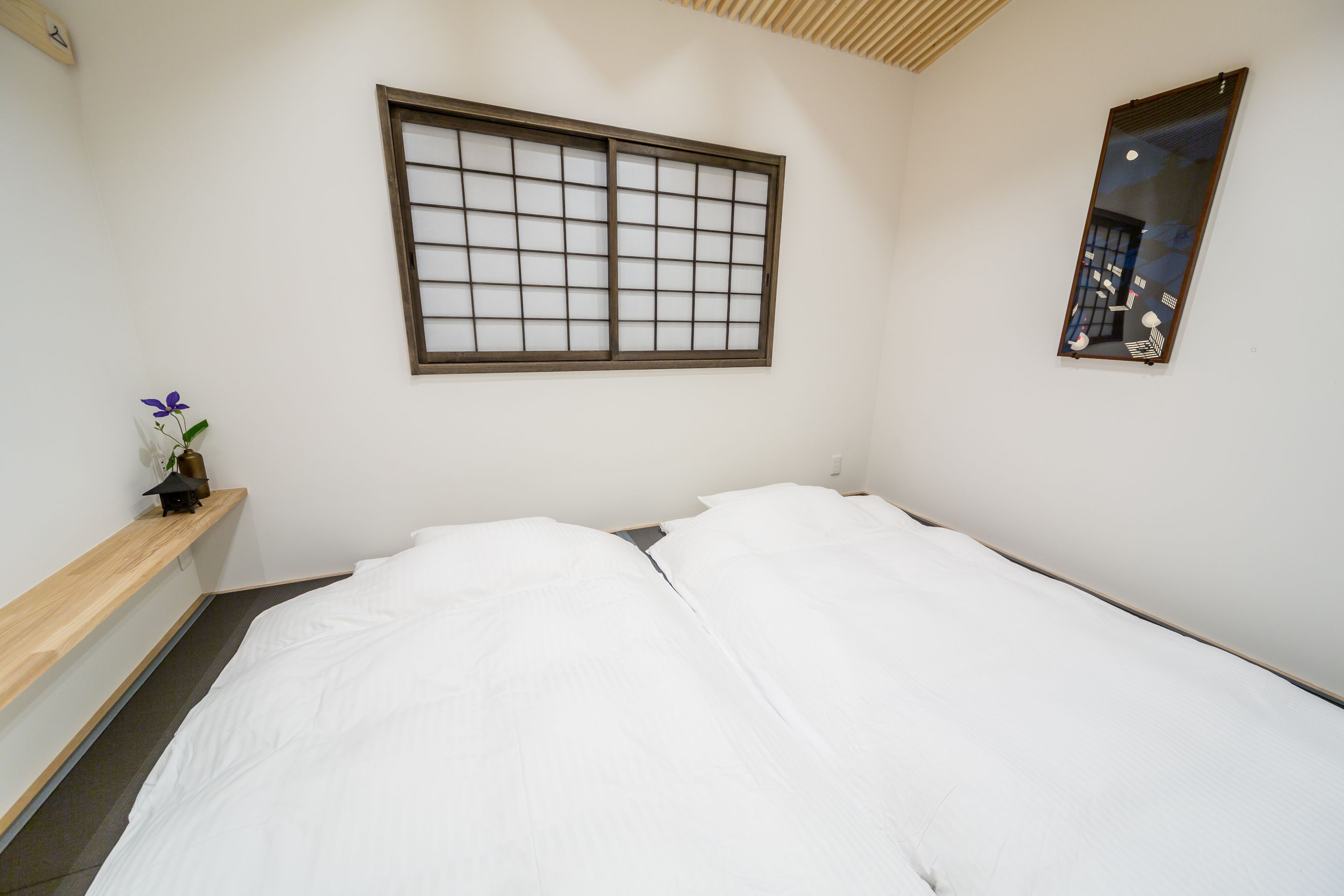 A room with a bed, a window with a grid pattern, a mirror, and a small shelf with a flower.