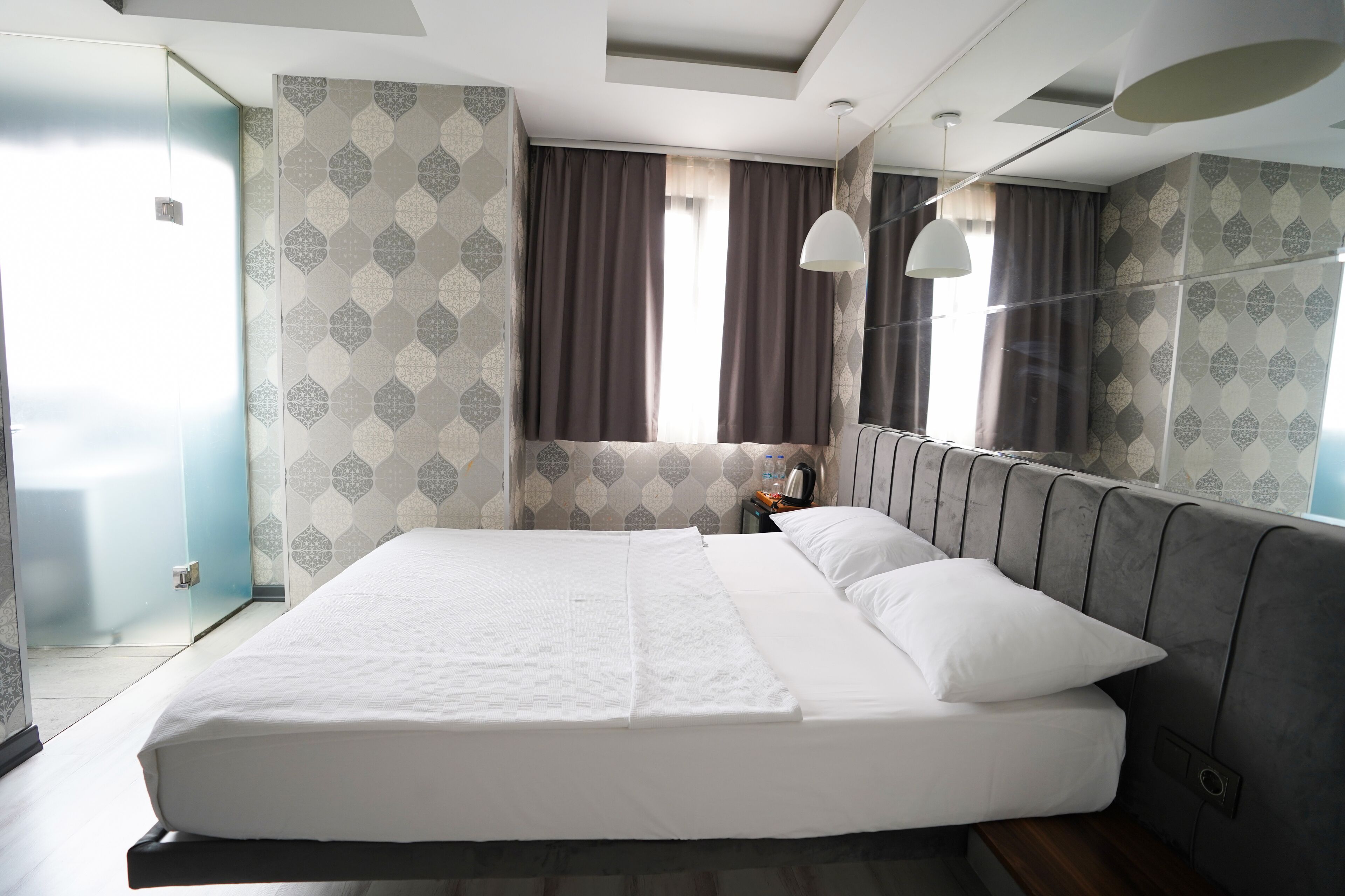 A modern hotel room with a large bed, a bedside table with a lamp, and patterned wallpaper.