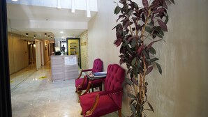 Um saguão de hotel com balcão de recepção, área de estar com poltronas vermelhas e uma grande planta em vaso.