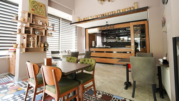 Uma sala de jantar com mesa e cadeiras de madeira, chão estampado e cozinha ao fundo.