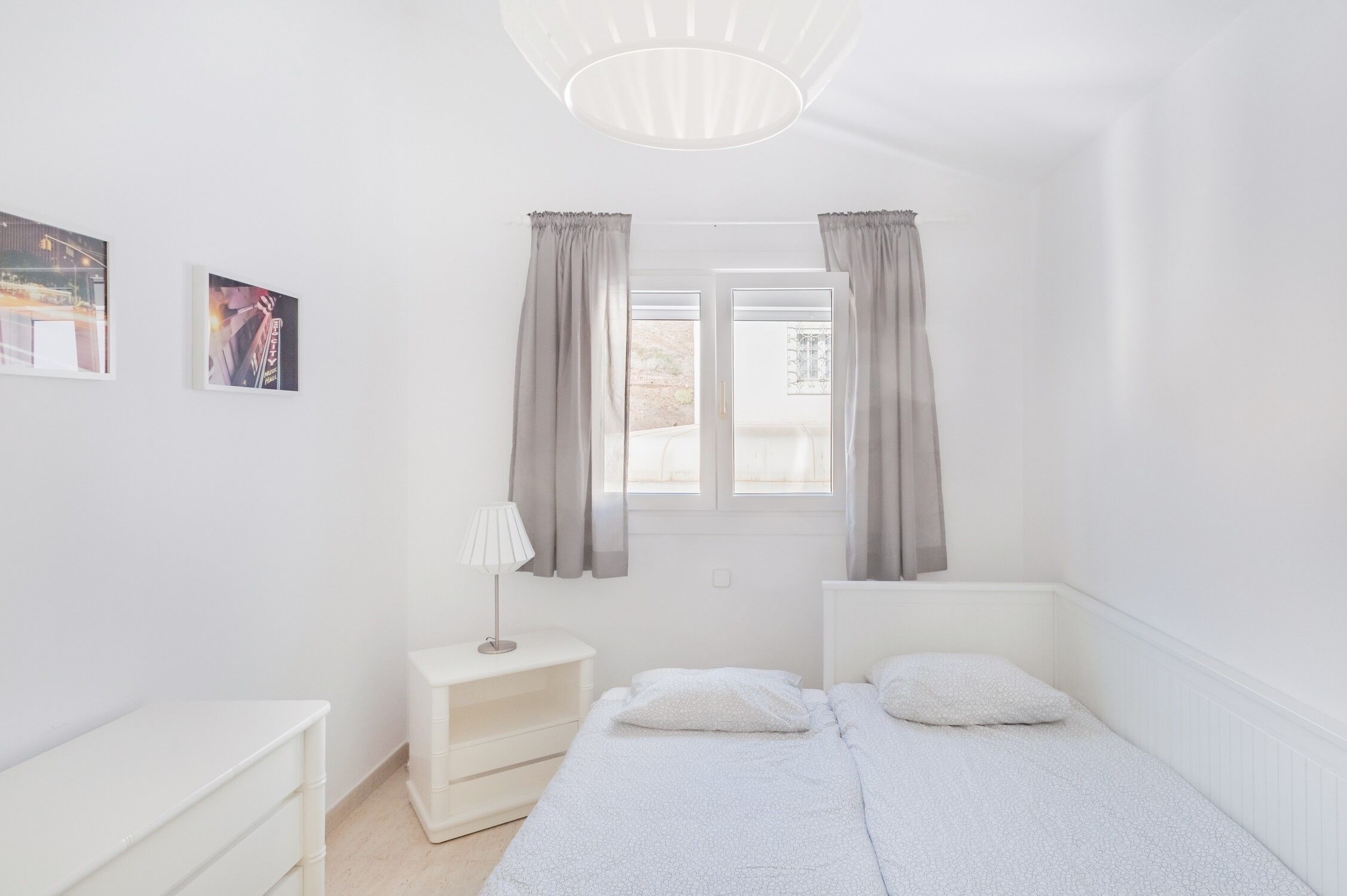 Minimalistinen makuuhuone, jossa on sänky, yöpöydät, lamppu ja kaksi ikkunaa, joissa on verhot.