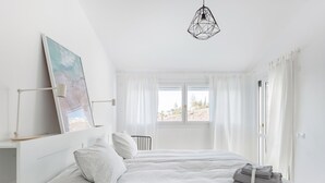 Minimalistinen makuuhuone, jossa on suuri sänky, yöpöytä, riippuvalaisin ja ikkuna, jossa on verhot.