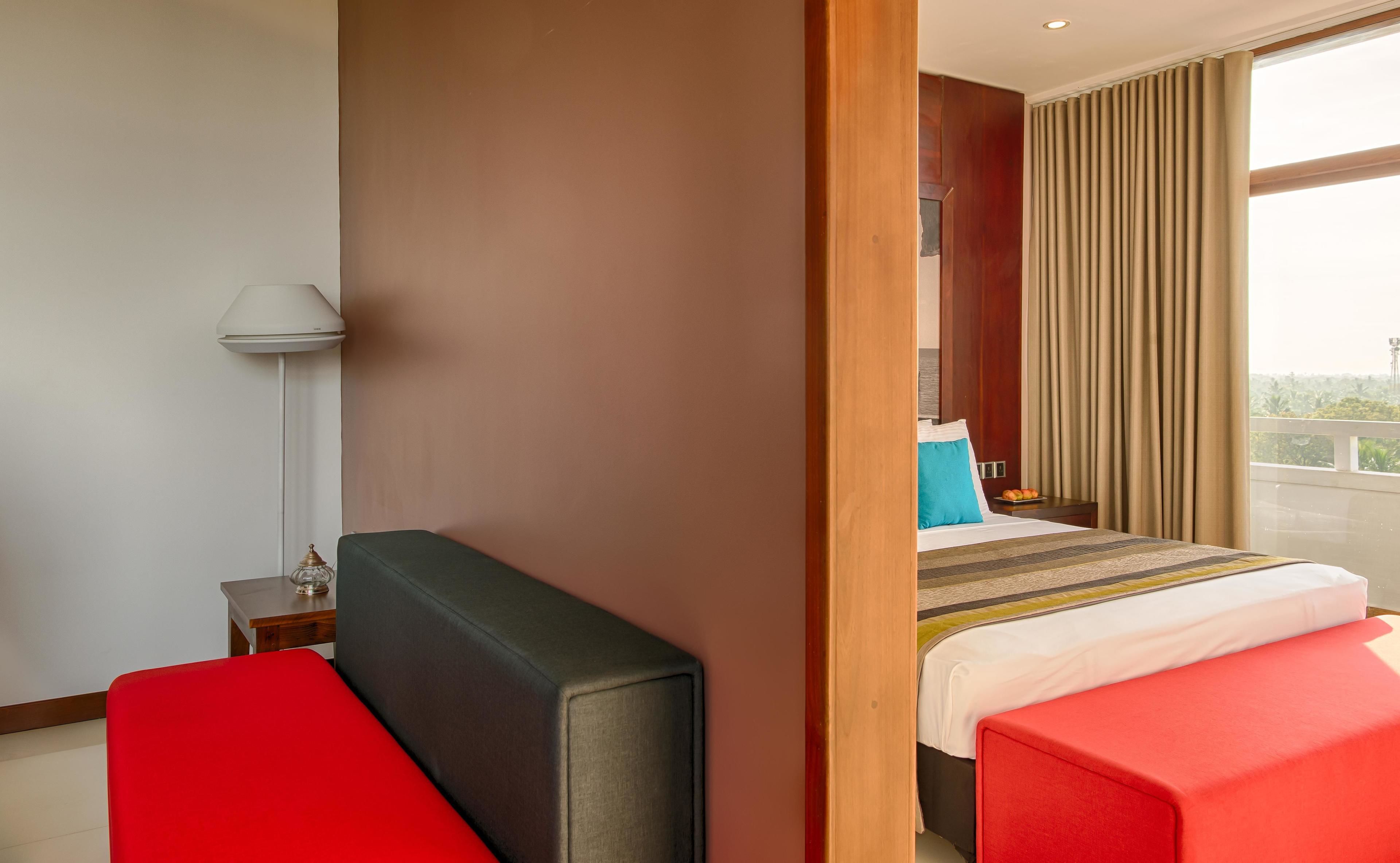 Une chambre d’hôtel avec un lit, une table de chevet, une lampe, un banc rouge et une vue sur l’extérieur.