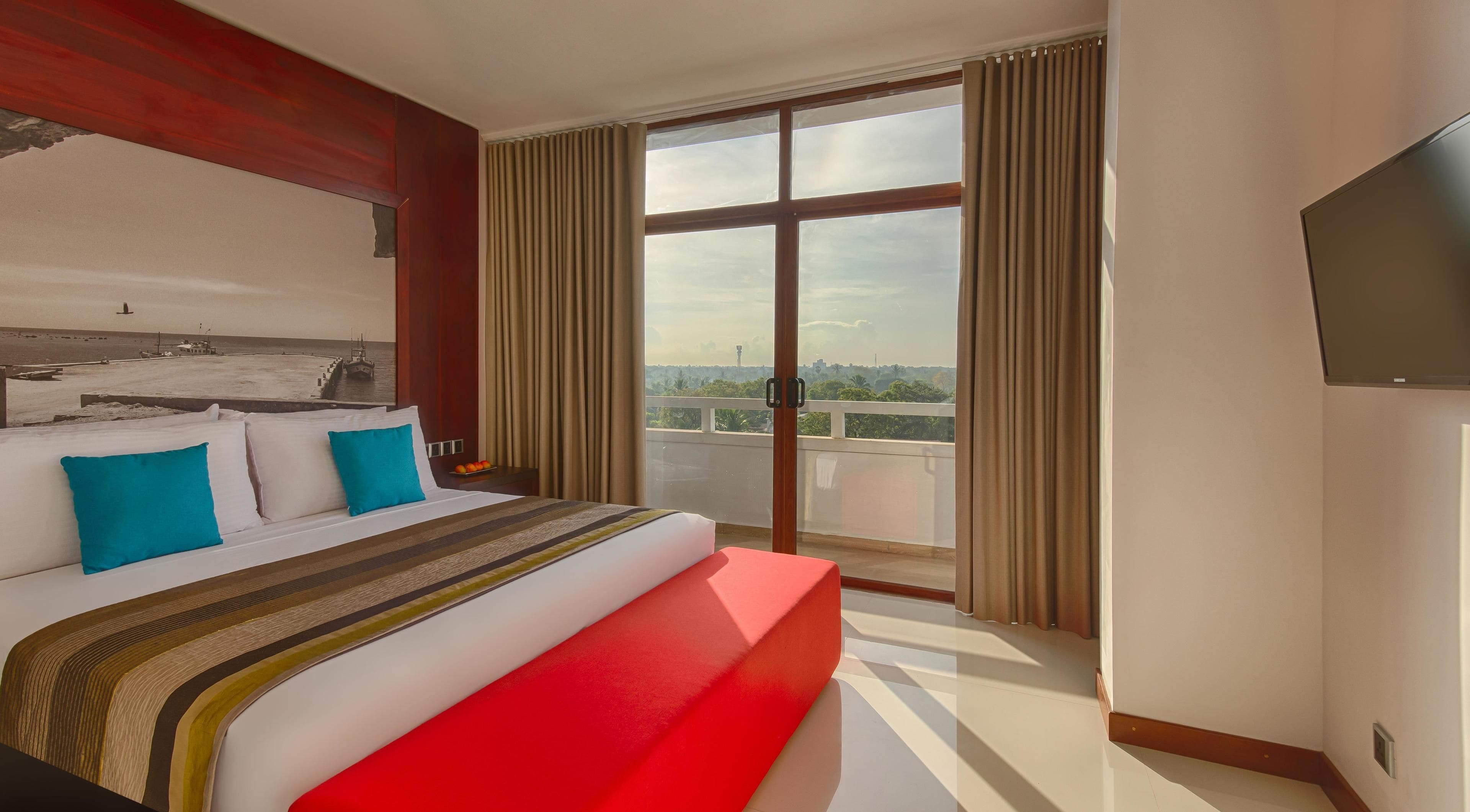 A hotel room with a large bed, a red accent wall, a balcony with a view, and a flat-screen TV.