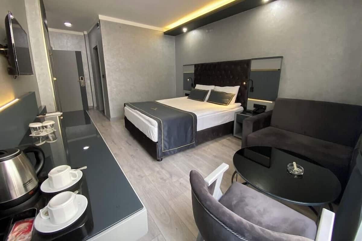 A modern hotel room with a bed, bedside tables, a desk with a coffee maker, a chair, and a small table.