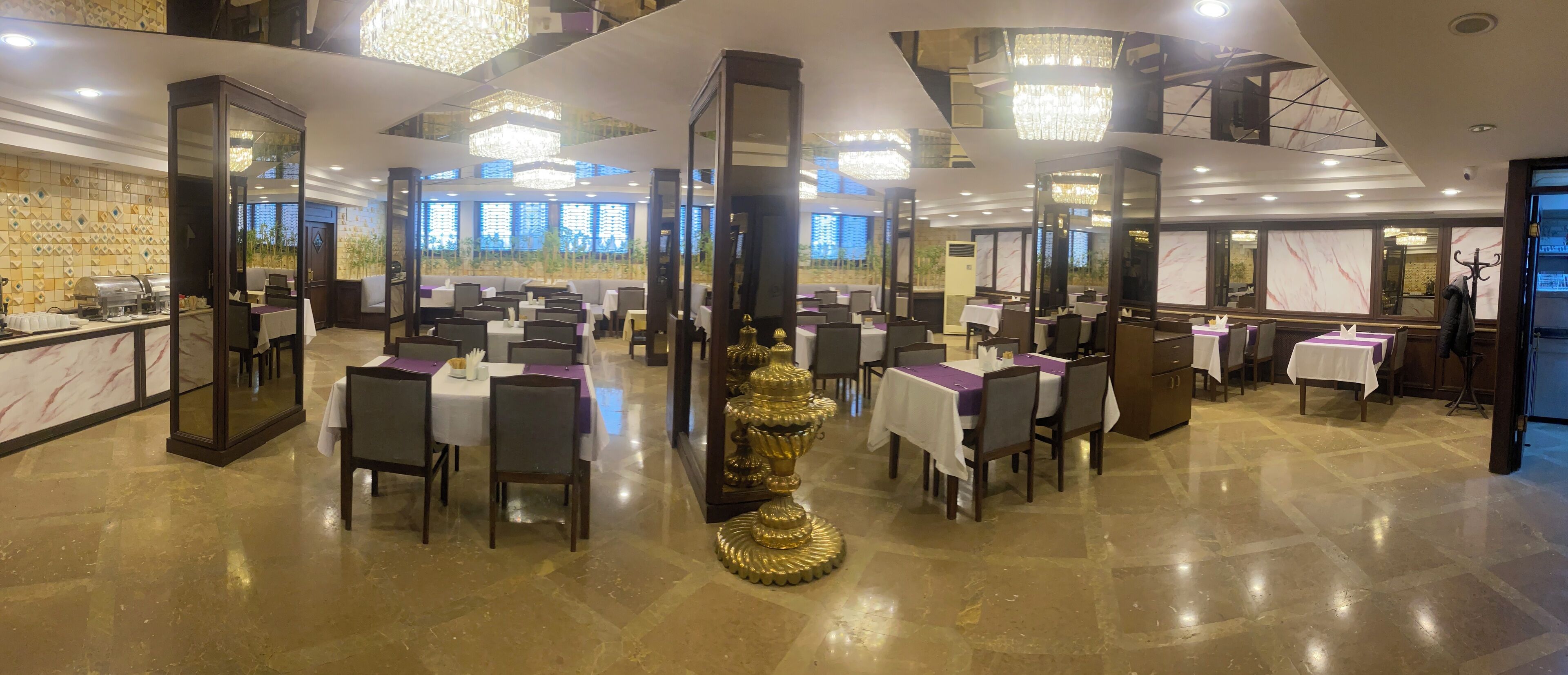 A spacious restaurant with multiple tables and chairs, a central decorative golden urn, and a modern chandelier.