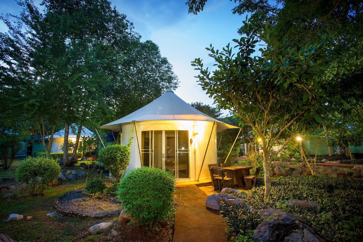 Een koepelvormige tent met een glazen deur, omgeven door een tuin met rotsen, struiken en bomen.