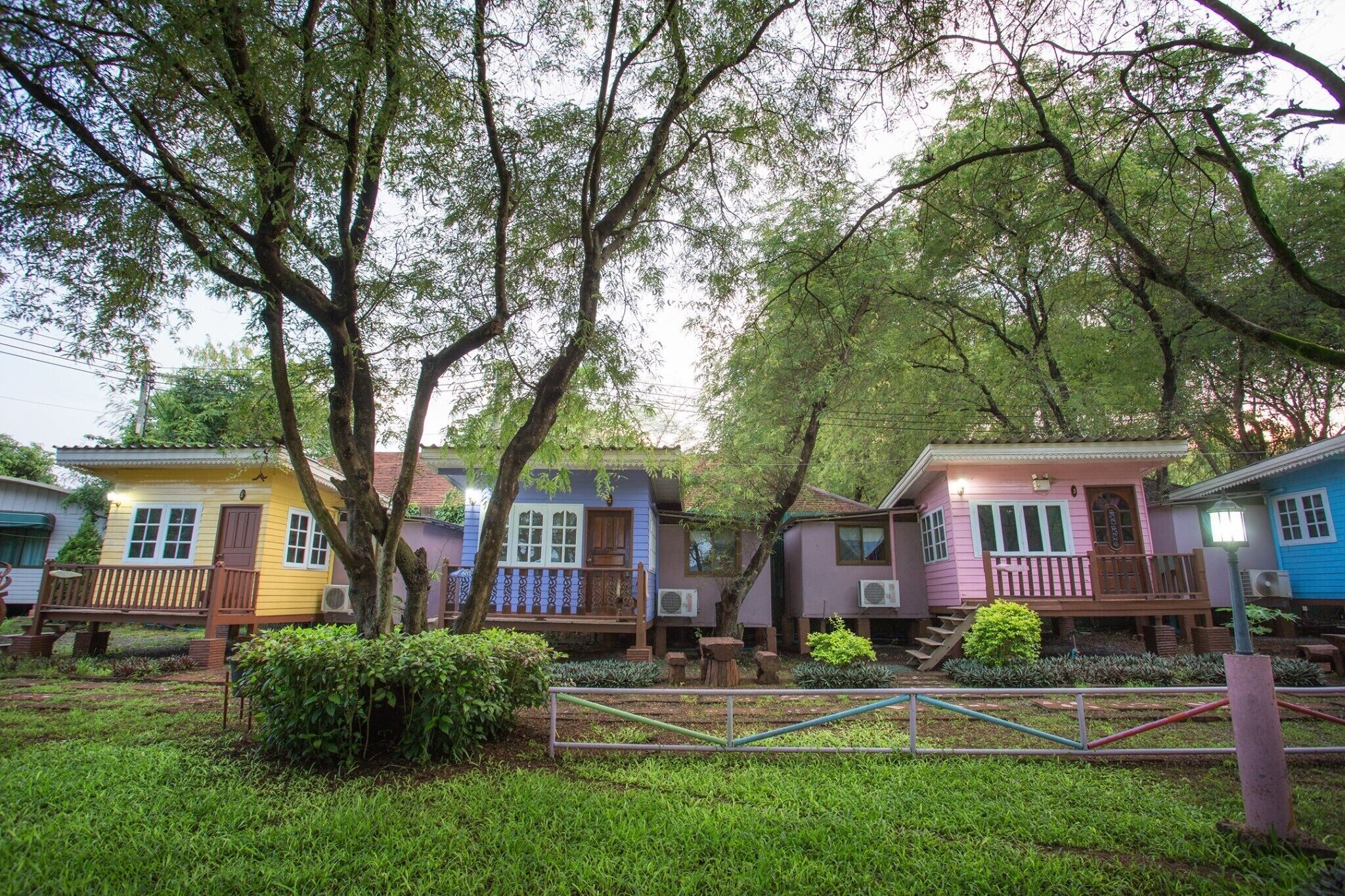 A row of colorful cabins with wooden decks and air conditioning units, surrounded by lush greenery and trees.