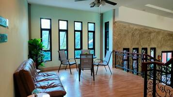 A spacious living area with a brown leather sofa, a small round table, and a dining table with chairs. Large windows provide natural light, and a balcony with a wrought iron railing is visible.