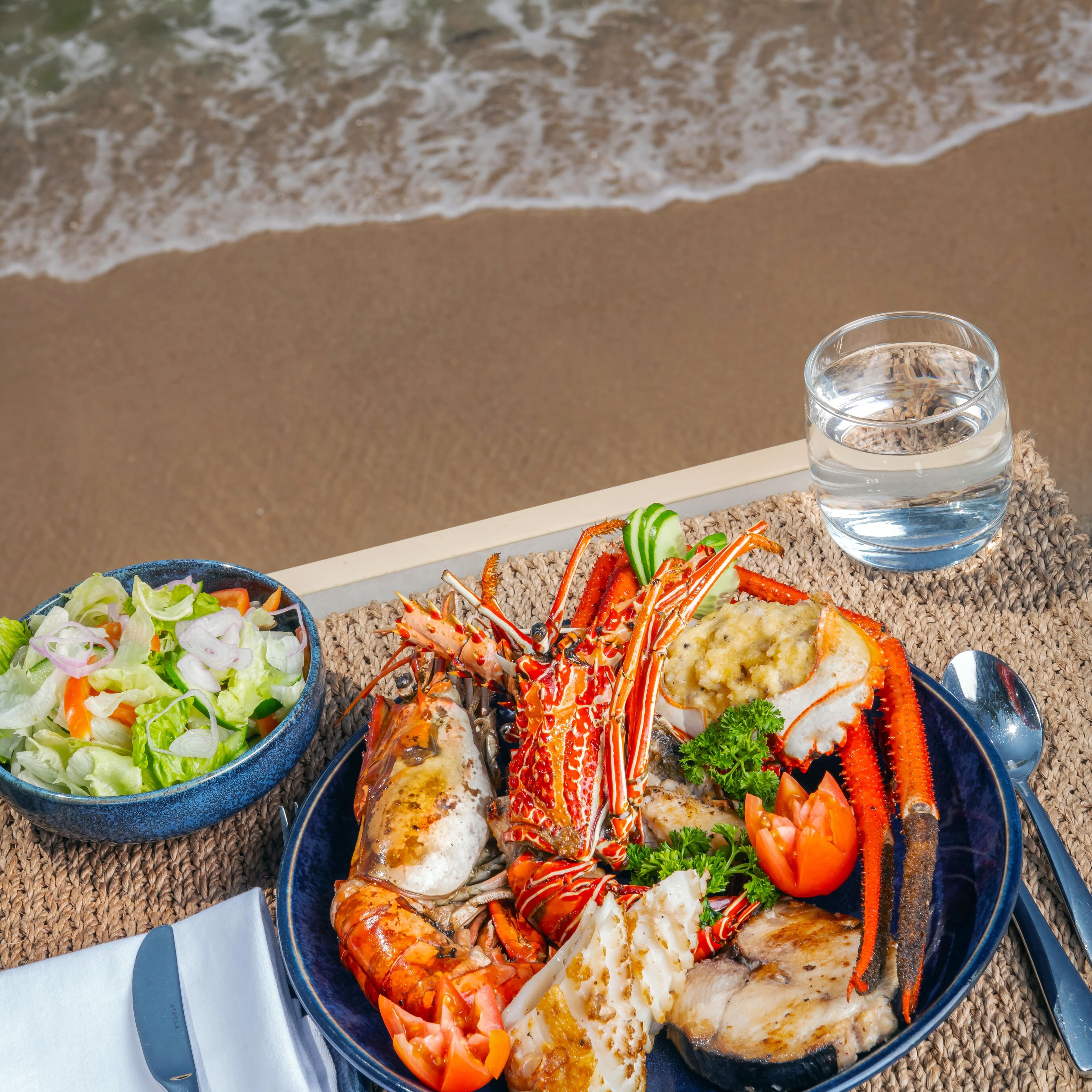 Uma refeição de frutos do mar sobre um tapete trançado à beira-mar, com um copo de água.