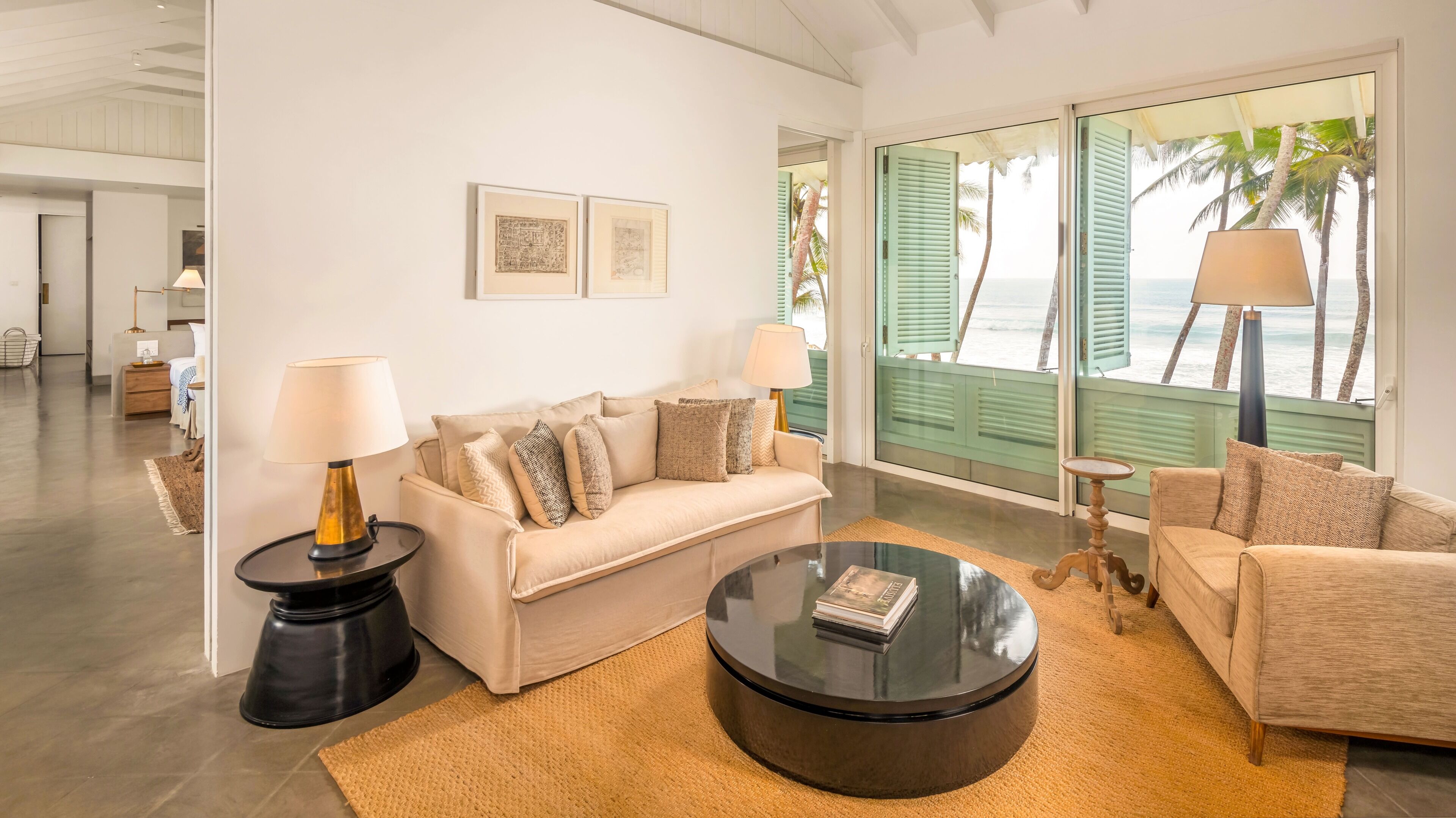 Una sala de estar moderna con un sofá color crema, dos mesitas de noche con lámparas, una mesa de centro y vistas al océano a través de ventanas grandes con contraventanas.