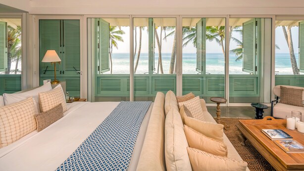 Quarto com cama, sofá, mesa de centro e vista para a praia através de venezianas abertas.