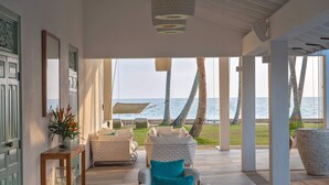 Amplo espaço interno com vista para o mar, com área de estar com móveis brancos e azuis, mesa de madeira com um vaso de flores e um quadro na parede.