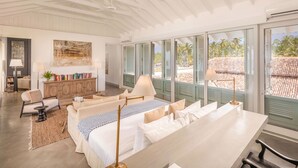 Quarto espaçoso com cama grande, mesa de jantar e vista para uma praia através de amplas janelas.