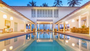 Uma área de piscina com vista clara para o oceano, um espaço de estar coberto e um edifício com janelas amplas e varanda.