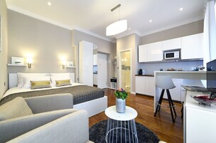 Una habitación de hotel moderna con cama, sofá, una pequeña mesa redonda con un jarrón, una zona de cocina con microondas y una mesa de comedor con sillas.