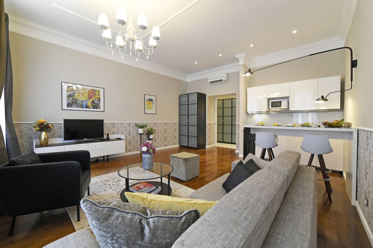 A modern living room with a sofa, coffee table, and TV. There is a kitchen area with a microwave and cabinets.