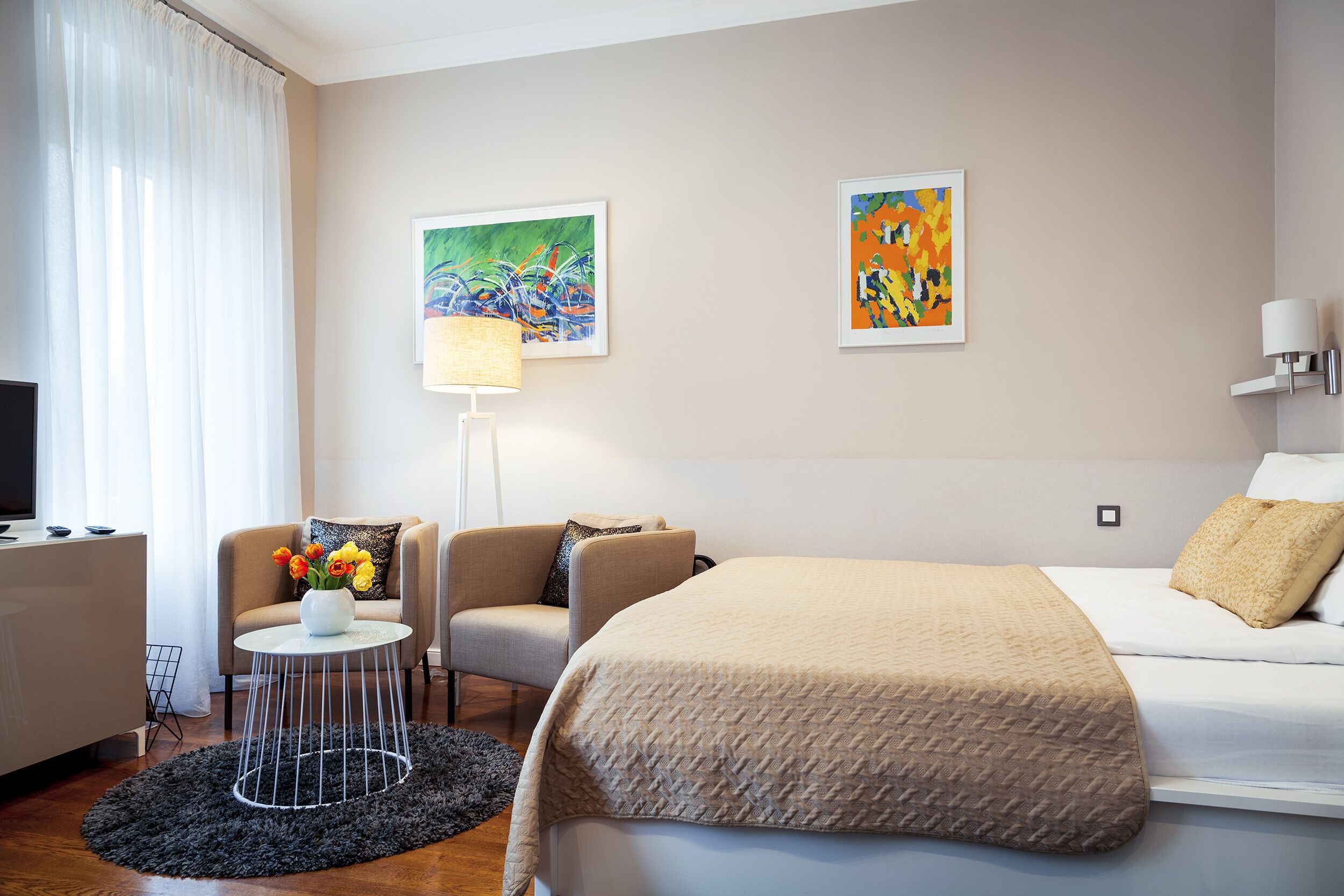 Camera d'albergo con un letto, un divano, un piccolo tavolino con un vaso di fiori, due quadri appesi al muro e una lampada.