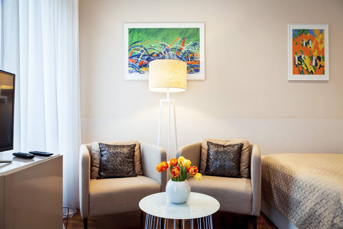 A hotel room with a bed, two armchairs, a small table with a vase of flowers, a floor lamp, and two paintings on the wall.