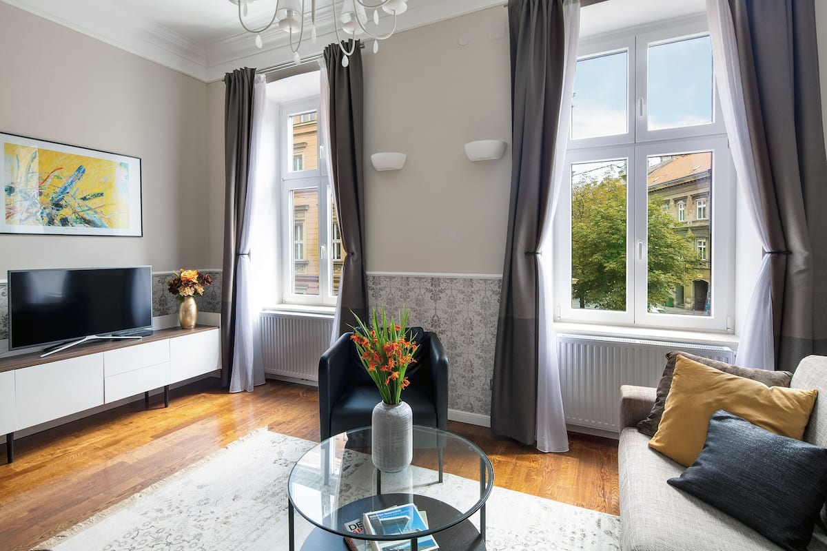 A modern living room with a flat-screen TV, a glass coffee table with a vase of flowers, a sofa with cushions, and large windows with curtains.