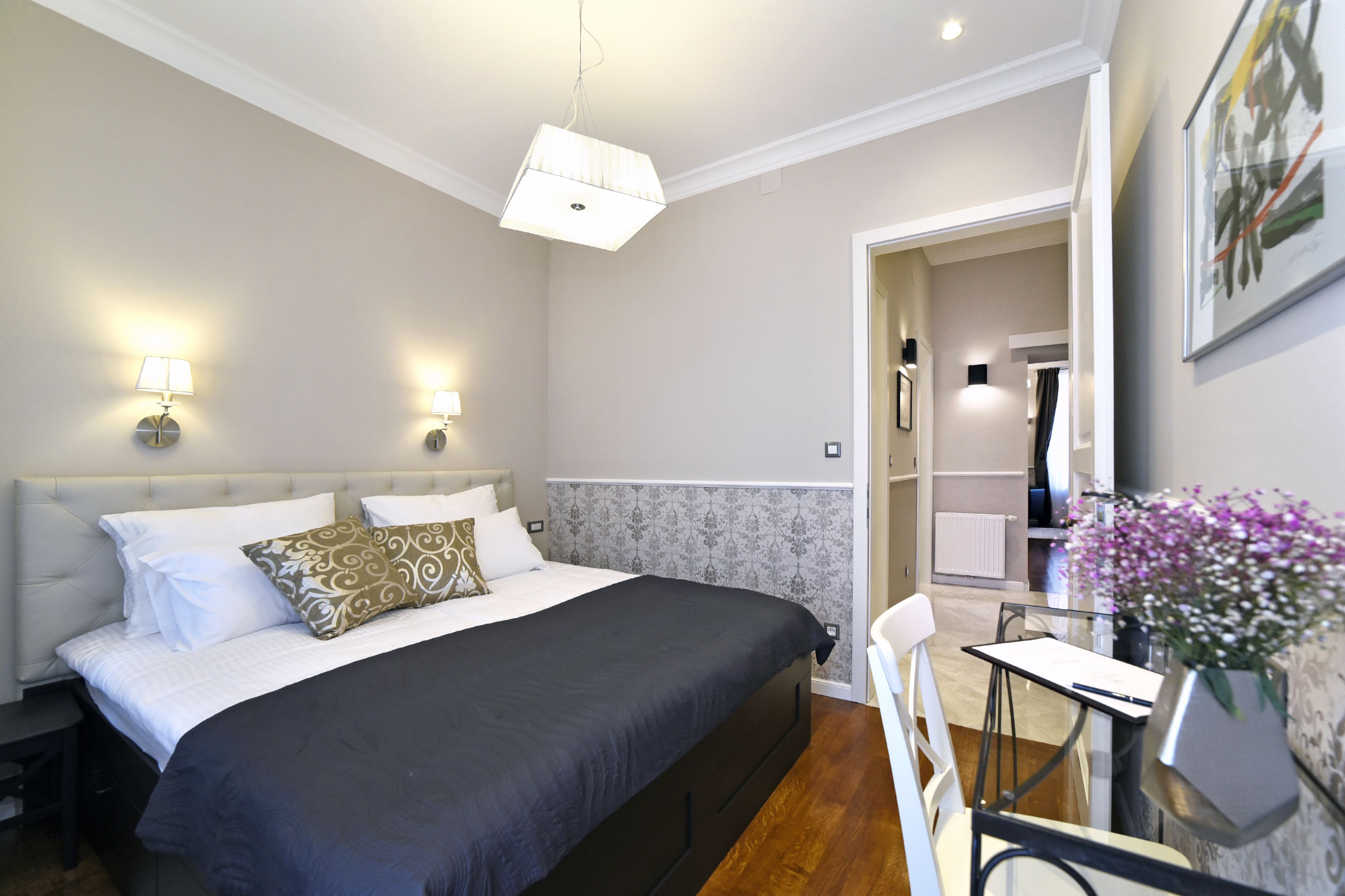 A bedroom with a large bed, bedside lamps, a desk with a chair, and a potted plant.