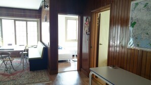 Una stanza con pavimento in legno, un tavolo bianco, una panca e una sedia. Si apre una porta verso un'altra stanza e c'è una finestra con persiane.